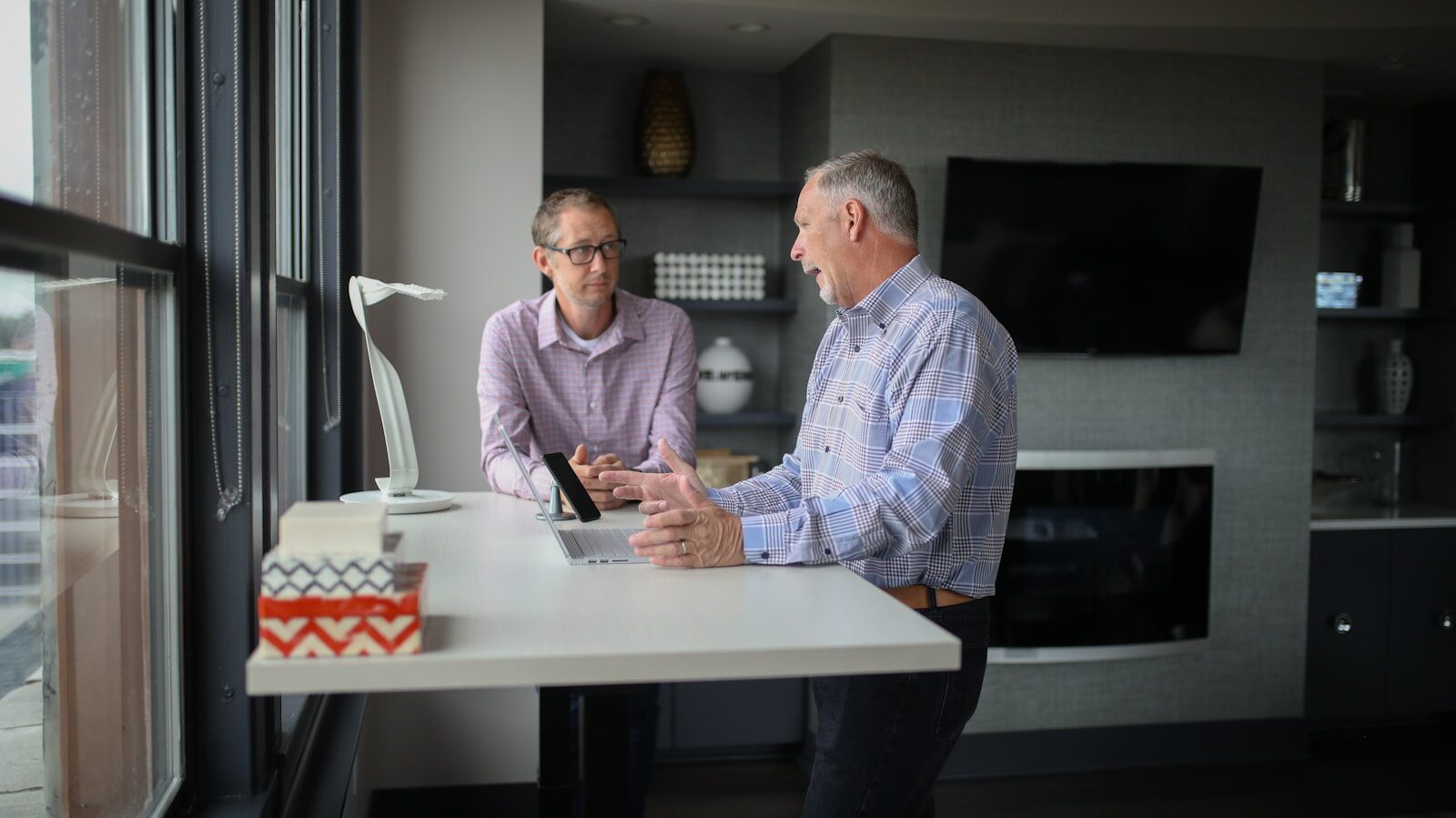 Two men engage in a conversation at a modern workspace, discussing ideas over a laptop. A desk lamp and decorative items are visible in the background.